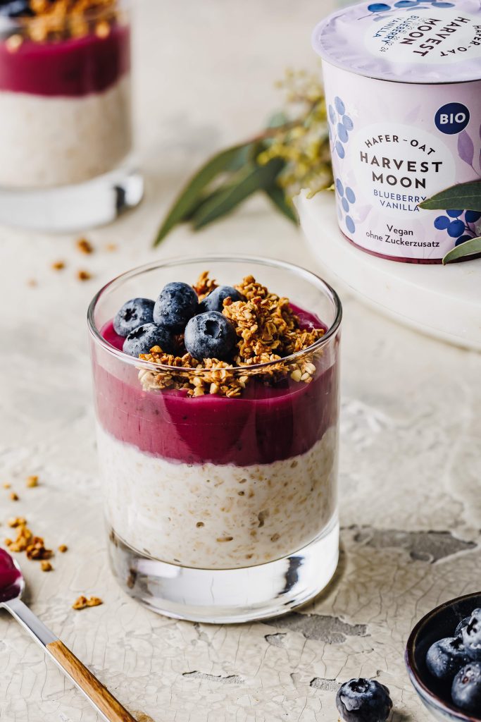 Auf diesem Produktfoto ist ein Heidelbeer Joghurt von Harvest Moon zu sehen. Im Vordergrund steht ein Glas mit Overnight Oats, dem Heidelbeer Joghurt und frischen Heidelbeeren.