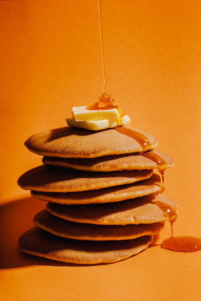 Hier sind saftige Pancakes auf orangem Hintergrund zu sehen, welche mit Honig übergossen werden.