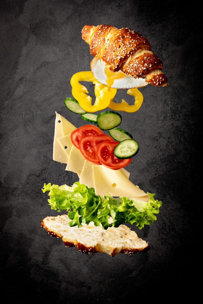 Hier ist ein Photoshop-Bild zu sehen von einem Croissant mit flying incredients. Jede einzelne Zutat des Food Fotos fliegt durch die Luft, wie das Croissant selbst, der Salat, der Käse, die Tomaten, die Gurken, der Paprika und das Spiegelei.