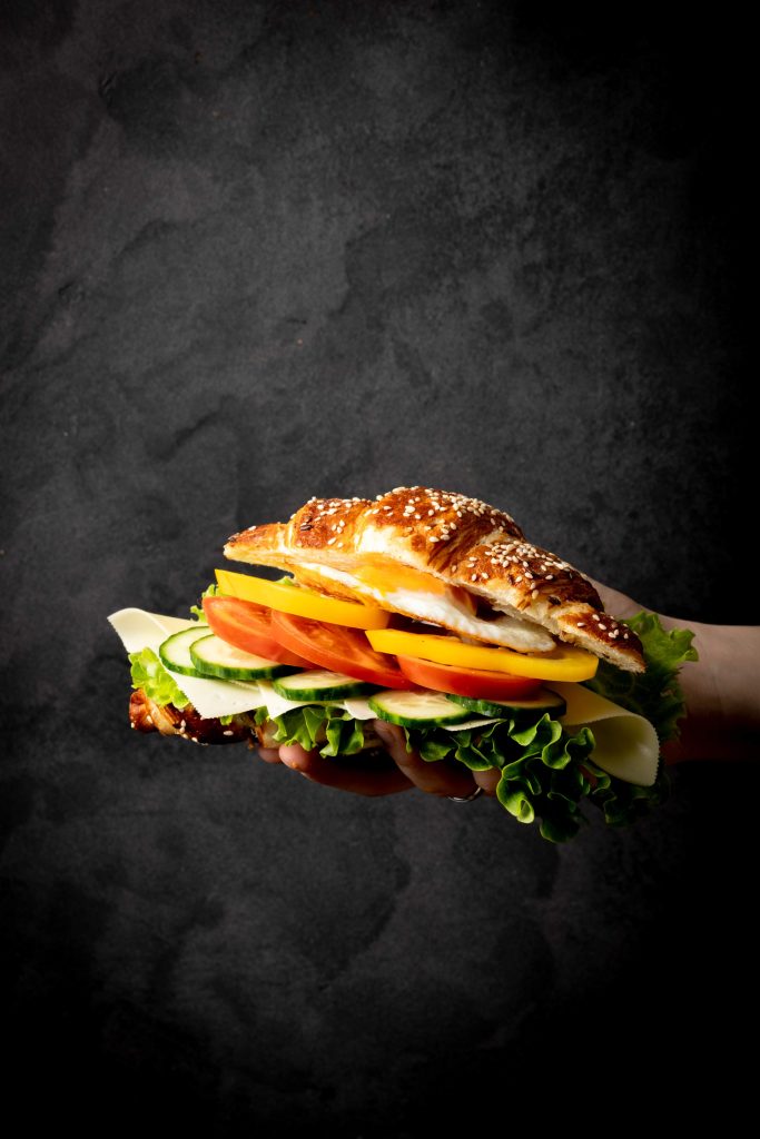 Auf diesem Food Foto ist ein Croissant vor einem schwarzen Hintergrund zu sehen. Das Croissant wurde mit Salat, Käse, Gurke, Tomaten, Paprika und Spiegelei belegt.