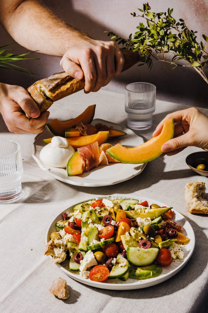 Dieses Bild zeigt eine große Tischszene. Auf dem Tisch steht ein Teller mit griechischem Salat, einer Zuckermelone, Prosciutto und Baguette.